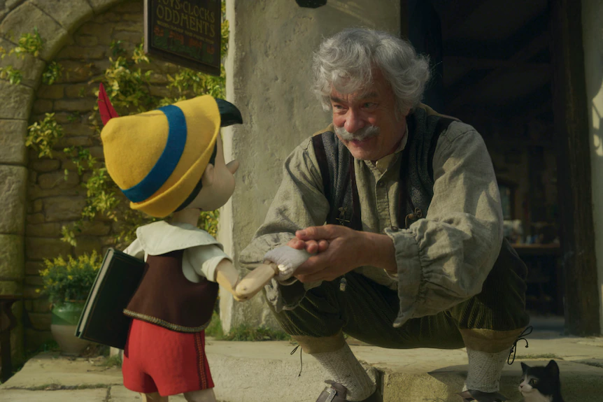 Vecchio con i capelli grigi inginocchiato e che interagisce con un piccolo giocattolo simile a un umano che indossa un cappello giallo, pantaloni rossi e un top bianco e marrone. 