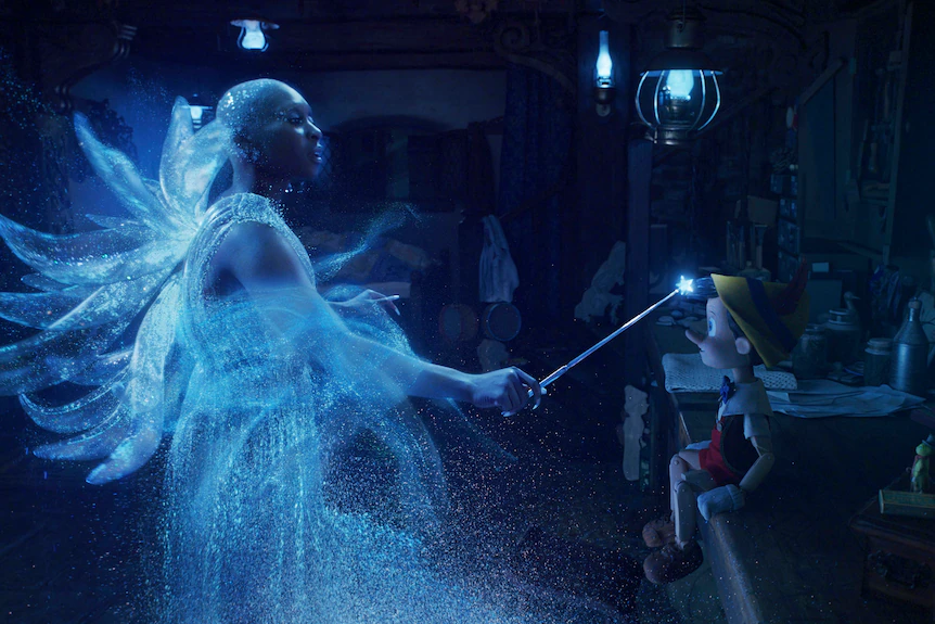 Una donna di colore in un costume da fata magica traslucido sta agitando una bacchetta contro un giocattolo in miniatura simile a un umano.  
